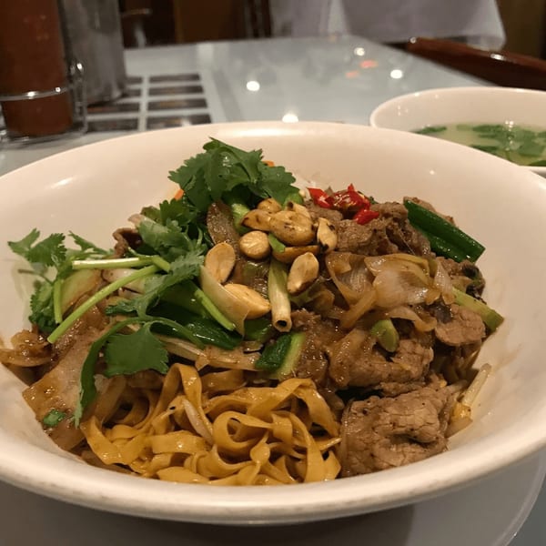A close-up of a Vietnamese noodle dish with beef, cilantro, peanuts, and chili in a white bowl.